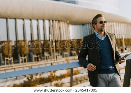A well-dressed man smiles while using a tablet on a balcony, surrounded by contemporary architecture and vibrant autumn foliage, enjoying a moment of leisure.