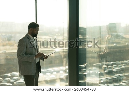 A well-dressed man smiles while using a tablet in a bright office space. The sunlight casts a warm glow, highlighting his focused expression as he enjoys a productive moment.