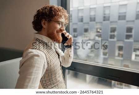 A welldressed man is smiling while engaging in a phone conversation inside a modern office environment