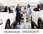 A well-dressed salesman is presenting information to Indian couple in a car dealership showroom. The couple appears interested as they stand between two new vehicles