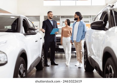 A well-dressed salesman is presenting information to Indian couple in a car dealership showroom. The couple appears interested as they stand between two new vehicles - Powered by Shutterstock