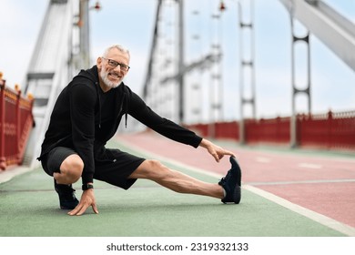 Well-fit athletic mature man in black sportswear and eyeglasses stretching legs while have workout outside on footbridge. Cheerful handsome elderly sportsman jogging by street, copy space - Powered by Shutterstock