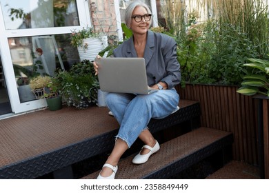 well-groomed senior woman with gray hair and glasses dressed in a gray jacket and jeans works remotely and walks along the street holding a laptop with her - Powered by Shutterstock