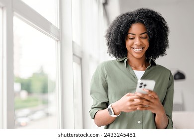 In a well-lit indoor setting, an African-American woman with a contagious smile interacts with her smartphone, engaging with friends or followers through a social network. - Powered by Shutterstock