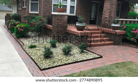 Well-maintained residential front yard featuring decorative landscaping with white river rocks, brick paver walkway, brick stairs, flowering shrubs, potted plants, and classic brick home entrance. Attractive suburban home exterior showcasing hardscape and low-maintenance garden design.