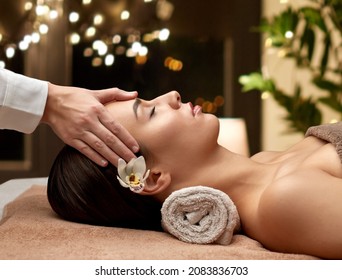 wellness, beauty and relaxation concept - beautiful young woman lying with closed eyes and having face and head massage at spa over christmas lights on window background
