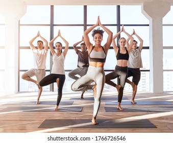 Wellness Concept. Multiethnic Group Of Sporty People Practicing Yoga With Instructor In Class, Standing In Tree Pose Against Window In Loft Studio