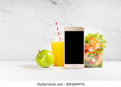 Wellness Fresh Summer Food - Set Of Orange Juice, Tropical Shrimp Salad In Plastic Box, Blank Screen Phone In White Bar Interior. Concept Advertising For Restaurant Take Away Or Delivery Service.