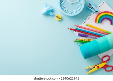 Well-organized drawing station from top view: bright stationery, pencil case, pens, sketchbook, plasticine, stapler, scissors, plane shaped sharpener, clock on soft blue backdrop. Space for text or ad