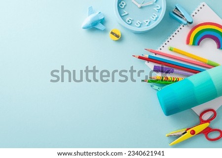 Well-organized drawing station from top view: bright stationery, pencil case, pens, sketchbook, plasticine, stapler, scissors, plane shaped sharpener, clock on soft blue backdrop. Space for text or ad