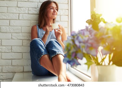 A well-pleased young girl enjoying an ideal day sitting in the window-sill. Wearing casual denim jeans and loose light-blue blouse, crossing legs and looking through the window - Powered by Shutterstock