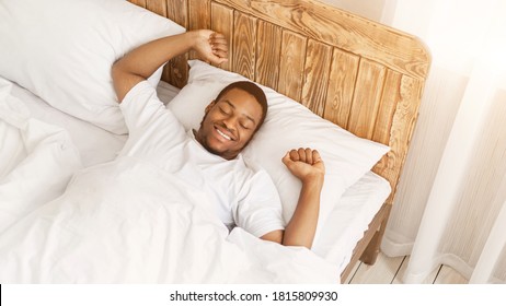 Wellslept African American Man Waking Up Stretching Hands Lying In Bed Smiling With Eyes Closed At Home. Awakening After Good Sleep In The Morning, Rest And Recreation Concept. High-Angle, Panorama