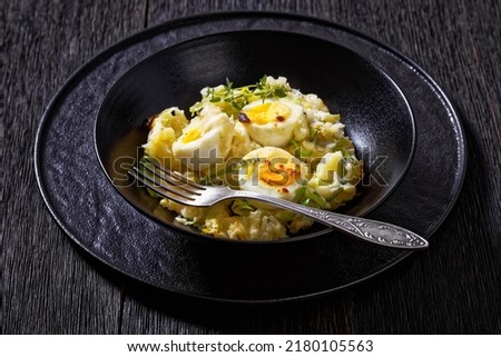 Welsh Anglesey Eggs, soft mashed potatoes with hard-boiled eggs all smothered in thick creamy leek and cheese sauce in black bowl on dark oak table, horizontal view
