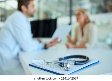 Were Always Here To Listen To Your Health Concerns. Shot Of A Stethoscope And A Clipboard On A Desk With A Doctor And Patient In The Background.