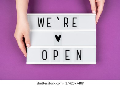 We're Open Text On A Lightbox On A Purple Background In Women's Hands. A Message From The Owner Of A Business, Cafe, Or Beauty Salon.