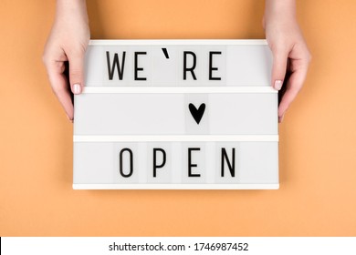 We're Open Text On A Lightbox On An Orange Background In Women's Hands. A Message From The Owner Of A Business, Cafe, Or Beauty Salon.