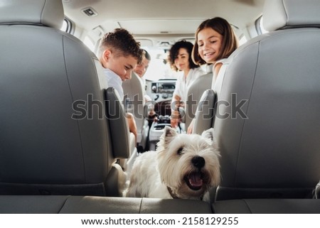 West Highland White Terrier Dog in Focus and Blurred Four Members Family Traveling by Minivan Car, Mother and Father with Two Teenage Children and Pet on Weekend Road Trip