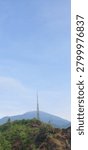 West Java, Indonesia - December 29, 2023 : Telecommunication tower lattice mast on a green hill with a majestic mountain and blue sky background.
In the background, a massive, serene mountain stands under a wide, clear blue sky with a few wisps of soft white clouds hugging its peak. The foreground highlights rocky terrain mixed with wild bushes and trees. This image perfectly illustrates telecommunications infrastructure, remote connectivity, network optimization, mobile coverage deployment in rural or mountainous areas, and engineering technology integrated with nature.