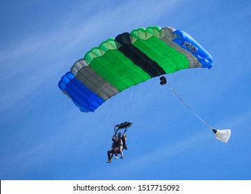 WEST PALM BEACH, FLORIDA - June 48, 2019: Tandem skydivers shown in silhouette as they near the ground underneath a blue and green parachute on a partly cloudy day