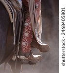 western cowboy boot with silver spur on cowgirl colored stylish boot with pattern and design on leather sitting on horse chinks rope and saddle pad visible close up vertical image room for type 