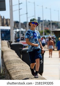 Weston Super Mare, Uk. July 21st. 2022. Energetic Boy Skipping Along Seaside Wall At Weston Promenade. Cheeky Young Boy, Facing The Camera Is Happily Singing And Playing. Dressed In Blue With Sun Hat.