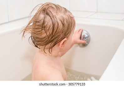 Wet Baby Girl With Blond Hair Looks At Her Reflection In Overflow Drain Knob Of The Bathtub.