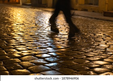 Wet Cobblestones At Night After Rain On A Street Or Road Surface, Bottom View With Shadow Or Silhouette Of Legs And Feet Of A Person Walking Or Running 