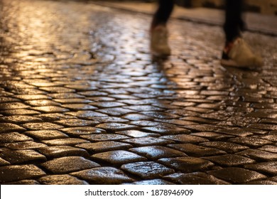 Wet Cobblestones At Night After Rain On A Street Or Road Surface, Bottom View With Shadow Or Silhouette Of Legs And Feet Of A Person Walking Or Running 