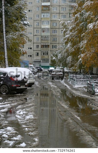 Wet road, sidewalk, iron black decorative\
fence covered with white fluffy snow, parked cars in the city yard.\
The first snow in autumn, in October. Trees with yellow leaves.\
Winter background