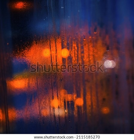 Wet steamy window inside bus on a rainy day, blurred orange and blue cars lights background - abstract moody background