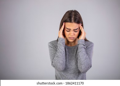 wful headache. oman holding head in hands and keeping eyes closed while standing against grey background. Woman suffering from headache, feeling strees. Studio shot, casual clothes, gray background.