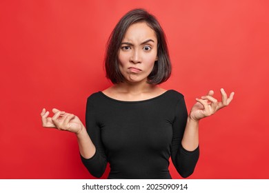 So What. Angry Asian Woman Raises Eyebrows From Annoynace Keeps Palms Sideways Shrugs Shoulders With Hesitation Wears Black Jumper Isolated Over Vivid Red Background Looks With Bewilderment.