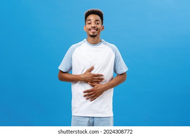 What Can Be More Satisfying Than Food. Delighted Good-looking Dark-skinned Guy With Afro Haircut Rubbing Belly Or Tummy And Smiling With Satisfaction Eating Delicious Tasty Food Over Blue Wall