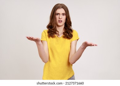 What do you want? Portrait of confused teenager girl in yellow T-shirt standing with raised hands and surprised indignant expression, asking what reason. Indoor studio shot isolated on gray background