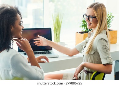 What do you think about it? Two smiling young women discussing something while sitting at working place 