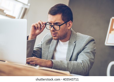 What is this? Handsome young man looking at his laptop and adjusting his glasses while sitting at his working place - Powered by Shutterstock