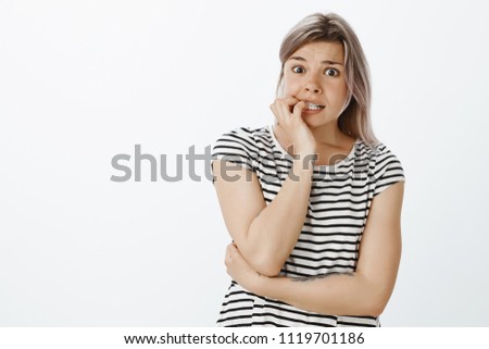 What if he knows it was me. Shocked nervous girlfriend in striped t-shirt, biting fingernails and gazing with scared expression at camera, feeling anxious and scared to answer for her mistakes