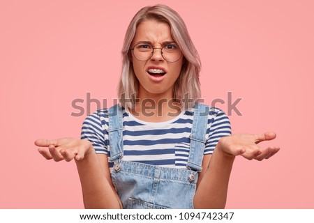 What hell are you talking about? Angry Caucasian young female keeps palms raised, being confused with dumb question, wears round big spectacles, gestures with annoyance, says: It`s nonsense!