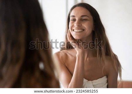 What a nice morning. Charming smiling young lady wrapped in big towel enjoying her reflection in bathroom mirror feeling happy having good mood satisfied with perfect healthy skin teeth hair condition