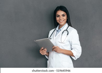 What Seems To Be A Problem? Attractive Young Female Doctor In White Lab Coat Holding Digital Tablet And Smiling While Standing Against Grey Background