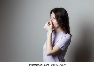 What A Smell. Close Up Of Young Unhappy Beautiful Woman With Long Hair In Casual T-shirt Squeezing Nose With Fingers, Disgust Expression, Feeling Bad About Bad Smell On Street