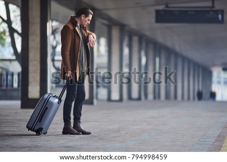 What time is it. Full length side view serious man looking at modern watch while keeping big suitcase. He waiting for train. Trip concept