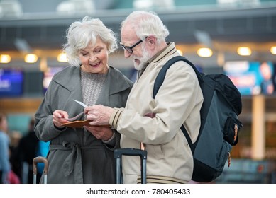 What Time Our Plane. Involved Gray-haired Couple Is Standing At Airport Terminal And Holding Flight Tickets. They Are Checking Travel Documents While Discussing Together With Smile