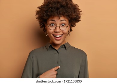 Am I what you need? Happy surprised female model points at herself, asks who me, smiles broadly, looks through round glasses, being chosen in business team, has positive reaction on hearing her name