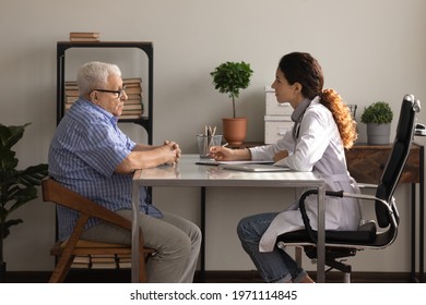 What Is Your Complaint. Side View Of Older Man Patient Meeting Young Woman Attending Physician For Regular Medical Checkup. Senior Male Clinic Visitor Talking With Latina Female Doctor At Appointment