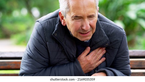 Whats Going On. Cropped Shot Of A Stressed Out Elderly Man Seated On A Bench And Holding His Chest In Discomfort Outside In A Park.