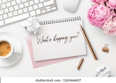 What's stopping you? Feminine workspace with bunch of peonies in a vase, computer, writing supplies, paper balls and a cup of coffee and hand-written quote on a white desk. Business concept. Flat lay.