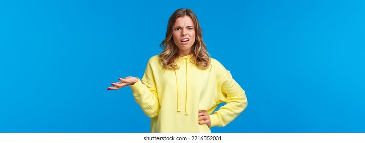 Whats wrong, why. Confused and frustrated blond girl dealing with person complains, shrugging and raise one hand in dismay grimacing perplexed, look camera questioned, blue background.