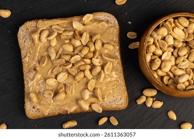 wheat bread with bran and soft peanut butter, peanut butter on a piece of bread and roasted peanuts on the table on black slate
