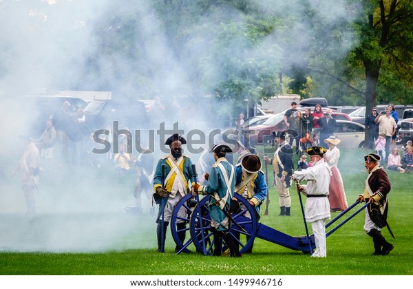 WHEATON,\
IL/USA - SEPT. 7, 2019: Gunsmoke drifts toward Continental forces\
preparing to move a cannon on a mock battlefield at a reenactment\
of the American Revolutionary War\
(1775-1783).